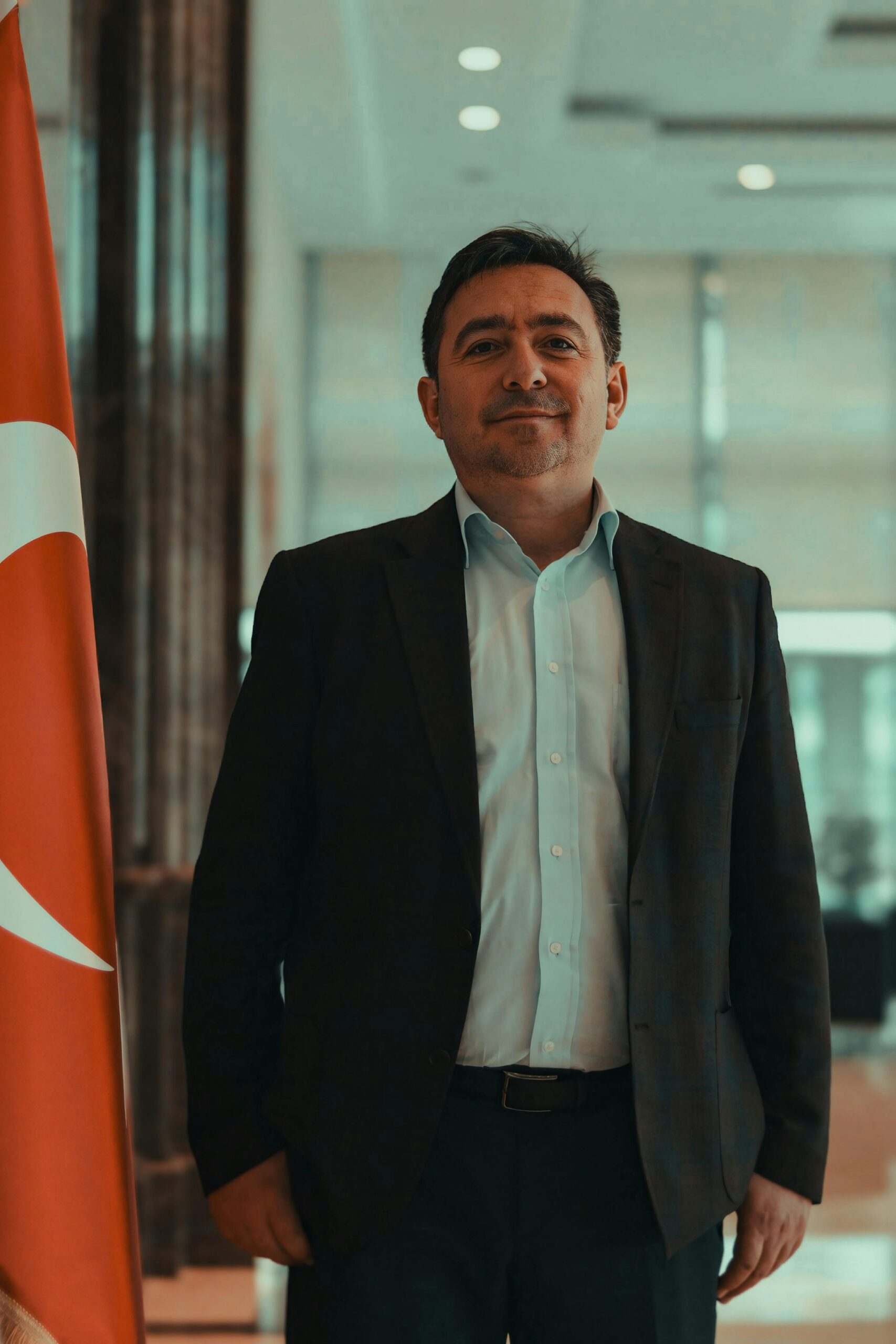 Middle-aged man in a suit standing confidently beside a Turkish flag indoors.