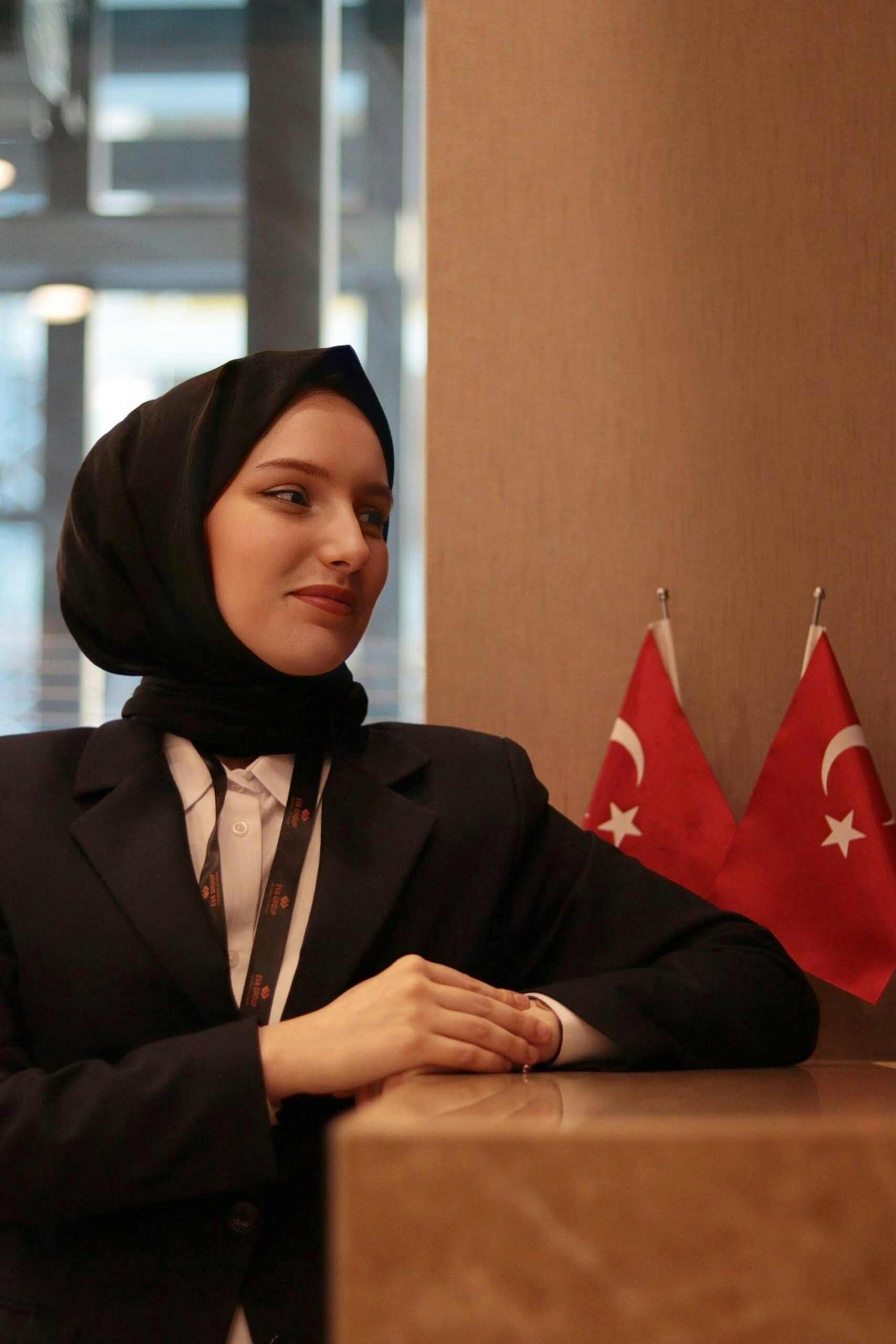 Portrait of a smiling woman in a black blazer with Turkish flags indoors.